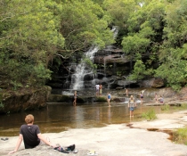 Person relaxing on rocks watching others in rockpool below waterfall
